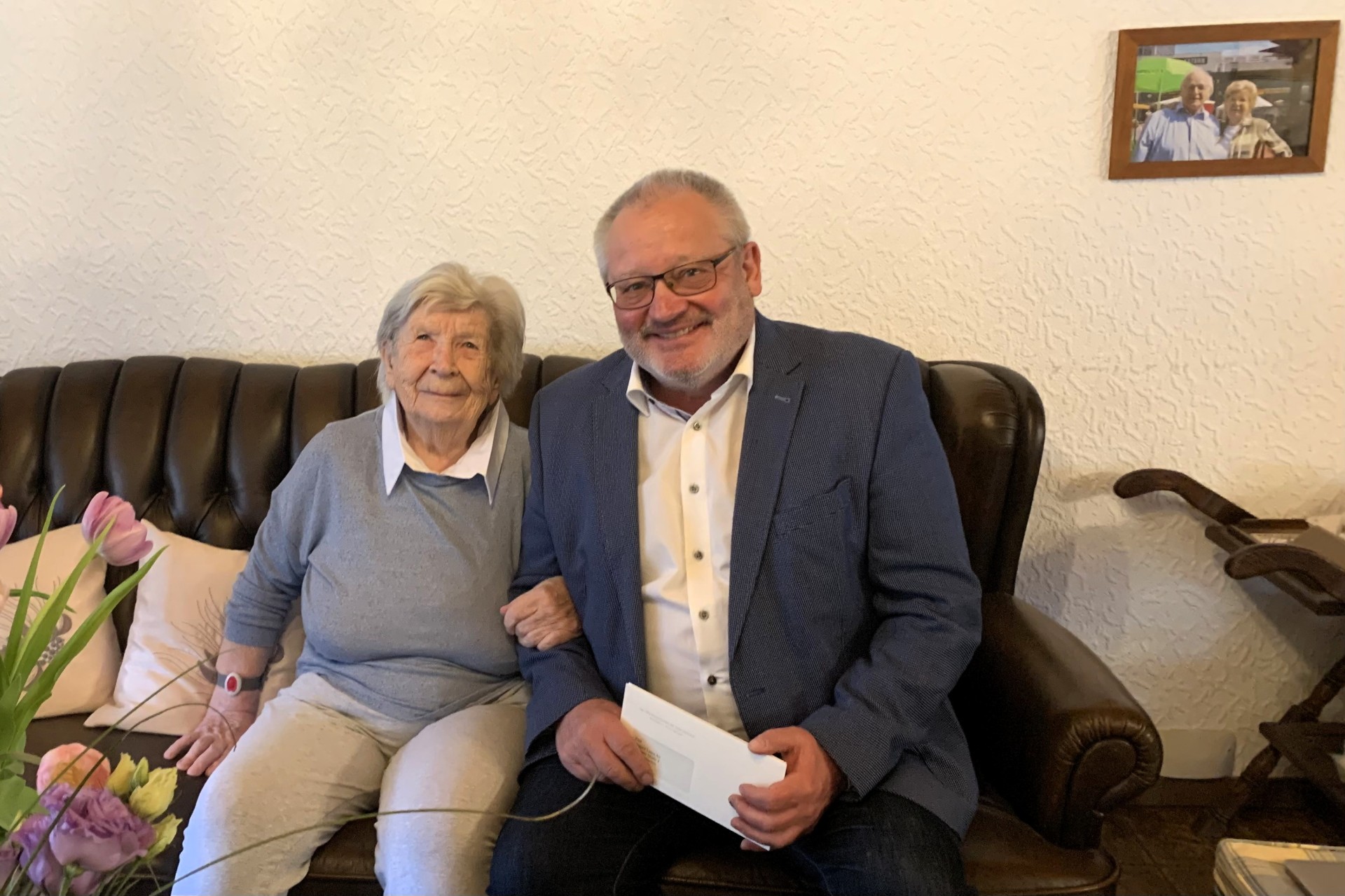 Eine ältere Dame sitzt lächelnd auf einem dunklen Ledersofa neben einem ebenfalls lächelnden Mann im Anzug. Die beiden sitzen nah beieinander, der Mann hält einen Briefumschlag in der Hand. Im Vordergrund ist ein Blumenstrauß zu sehen, im Hintergrund hängt ein gerahmtes Foto an der Wand. Die Szene wirkt freundlich und familiär.