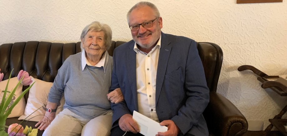 Eine ältere Dame sitzt lächelnd auf einem dunklen Ledersofa neben einem ebenfalls lächelnden Mann im Anzug. Die beiden sitzen nah beieinander, der Mann hält einen Briefumschlag in der Hand. Im Vordergrund ist ein Blumenstrauß zu sehen, im Hintergrund hängt ein gerahmtes Foto an der Wand. Die Szene wirkt freundlich und familiär.