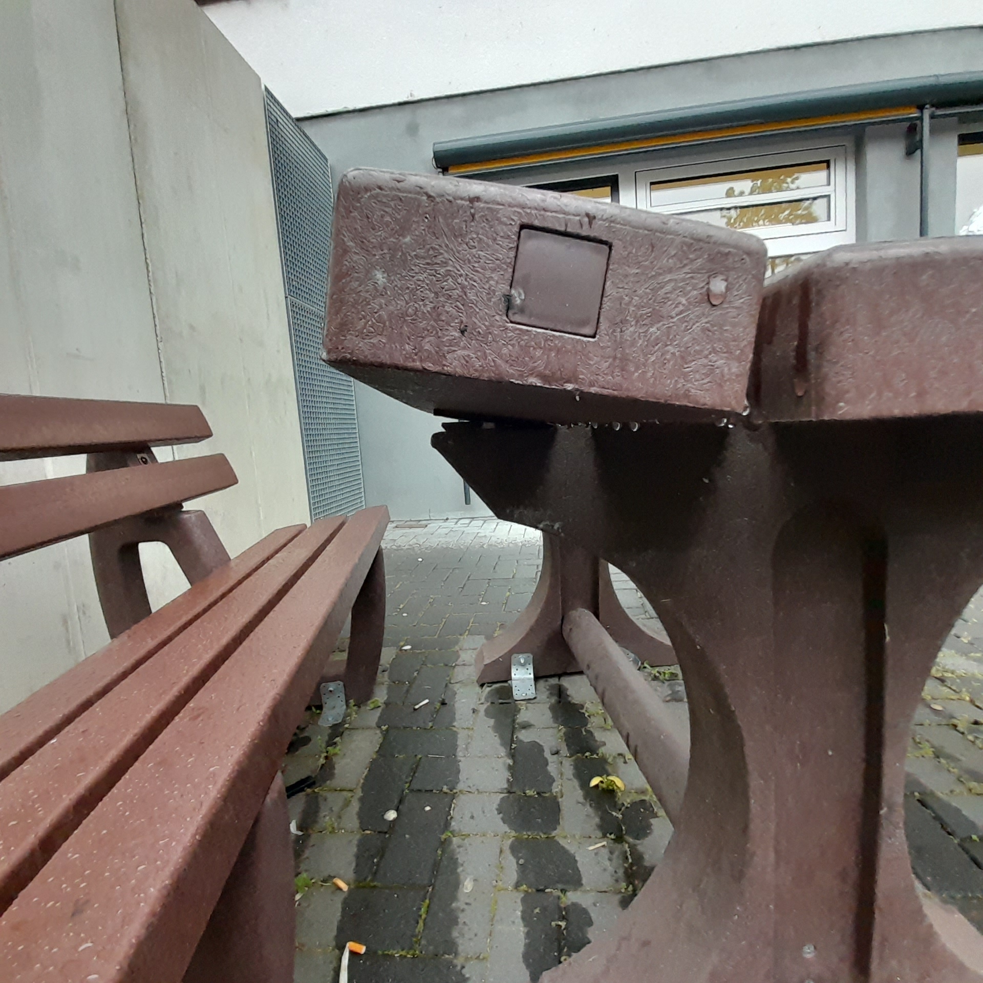 Beschädigte Sitzgruppe auf einem Schulhof: Ein schwerer Tisch aus braunem Material ist teilweise aus seiner Verankerung gerissen und steht schief, die Tischplatte wirkt beschädigt. Daneben befinden sich passende Bänke auf einem gepflasterten Boden, im Hintergrund ist ein Schulgebäude zu sehen.