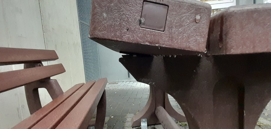 Beschädigte Sitzgruppe auf einem Schulhof: Ein schwerer Tisch aus braunem Material ist teilweise aus seiner Verankerung gerissen und steht schief, die Tischplatte wirkt beschädigt. Daneben befinden sich passende Bänke auf einem gepflasterten Boden, im Hintergrund ist ein Schulgebäude zu sehen.