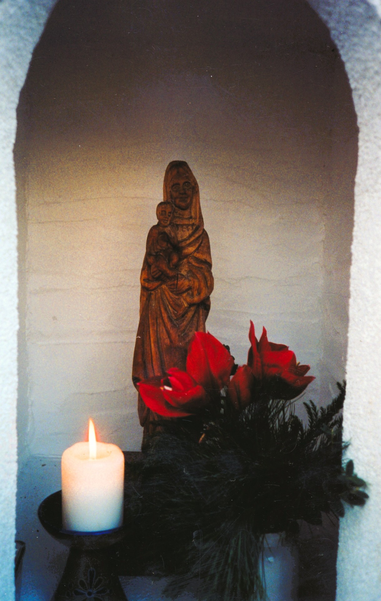 Holzfigur der Madonna mit Kind in einer kleinen, weißen Nische; davor stehen rote Blumen und eine brennende Kerze auf einem schwarzen Halter, die Szene wirkt ruhig und andächtig.