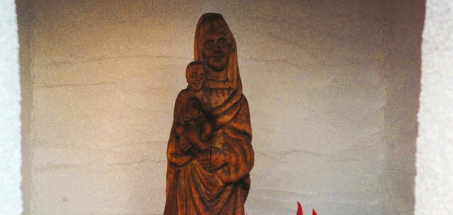 Holzfigur der Madonna mit Kind in einer kleinen, weißen Nische; davor stehen rote Blumen und eine brennende Kerze auf einem schwarzen Halter, die Szene wirkt ruhig und andächtig.