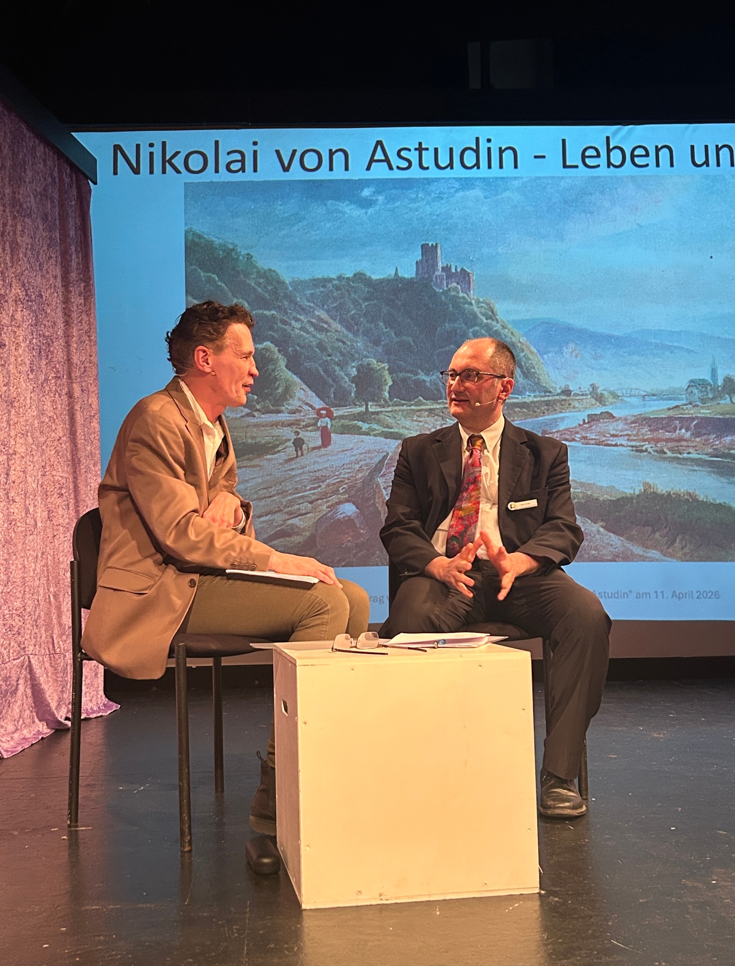 Zwei Männer sitzen auf einer Bühne im Gespräch, im Hintergrund eine projizierte Folie über Nikolai von Astudin mit einer Rheinlandschaft.