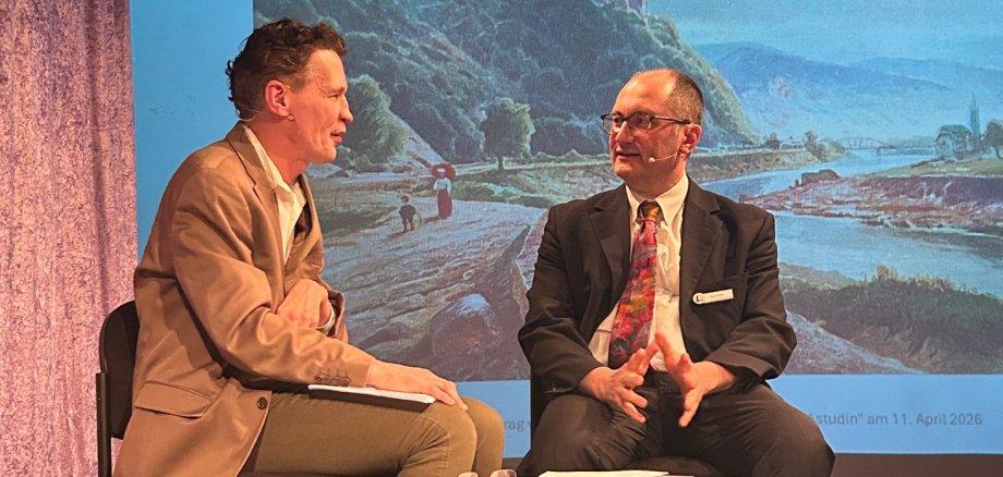 Zwei Männer sitzen auf einer Bühne im Gespräch, im Hintergrund eine projizierte Folie über Nikolai von Astudin mit einer Rheinlandschaft.