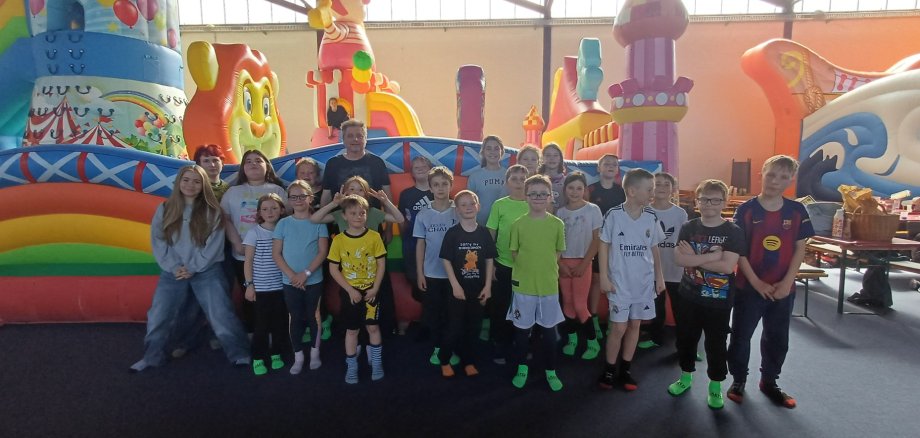 Eine Gruppe von Kindern und Betreuenden steht in einer Indoor-Spielhalle vor großen, bunten Hüpfburgen und schaut in die Kamera. Viele Kinder tragen bunte Kleidung und Socken, die Atmosphäre wirkt fröhlich und lebendig.