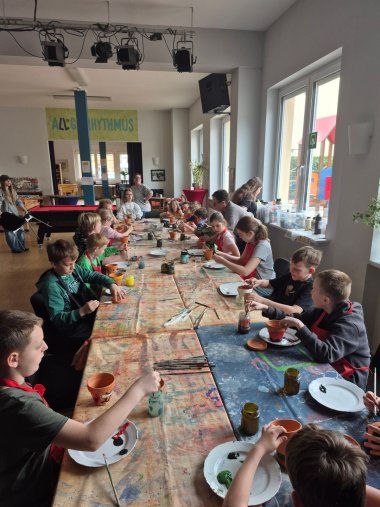 Mehrere Kinder sitzen an einem langen Tisch im Jugendkulturzentrum und bemalen kleine Terrakotta-Blumentöpfe mit Farben und Pinseln. Auf dem Tisch stehen Farbgläser und Teller, im Hintergrund ist ein heller Raum mit großen Fenstern zu sehen.