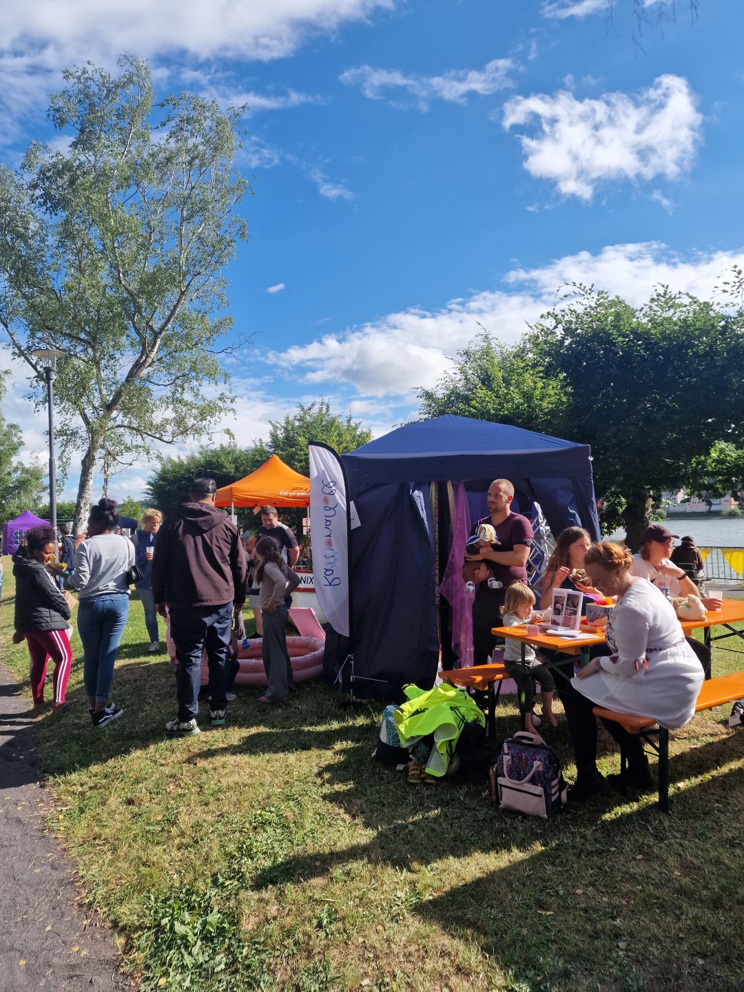 Mehrere Familien und Kinder stehen und sitzen bei sonnigem Wetter auf einer Wiese am Rhein. Im Vordergrund befindet sich ein Pavillon mit einem Stillzelt, daneben sitzen Erwachsene und Kinder an Bänken. Im Hintergrund sind weitere bunte Zelte und Besucher zu sehen.