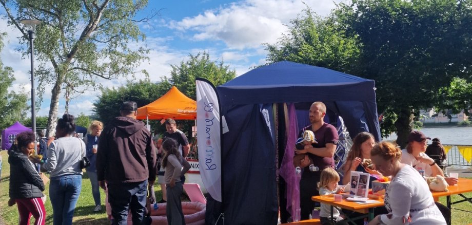 Mehrere Familien und Kinder stehen und sitzen bei sonnigem Wetter auf einer Wiese am Rhein. Im Vordergrund befindet sich ein Pavillon mit einem Stillzelt, daneben sitzen Erwachsene und Kinder an Bänken. Im Hintergrund sind weitere bunte Zelte und Besucher zu sehen.
