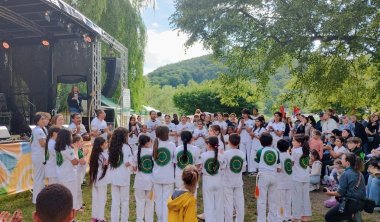 Eine Gruppe von Kindern und Jugendlichen in weißen Capoeira-Outfits steht im Kreis vor einer Bühne und klatscht. Zuschauerinnen und Zuschauer verfolgen die Aufführung im Freien unter Bäumen.