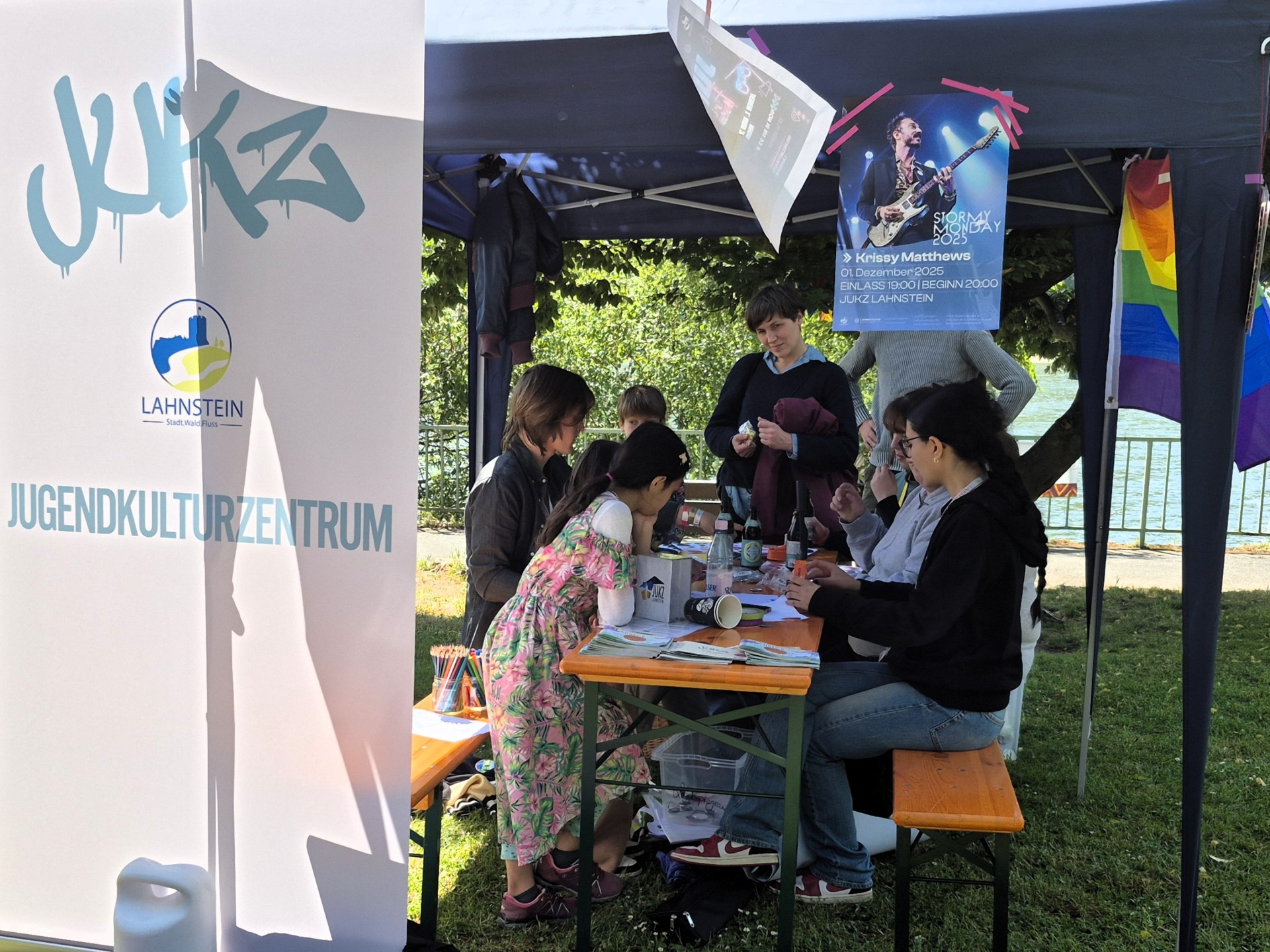 Unter einem Pavillon sitzen Kinder und Erwachsene an einem Tisch und basteln gemeinsam mit verschiedenen Materialien. Daneben steht ein Banner des Jugendkulturzentrums Lahnstein, im Hintergrund sind Bäume und der Rhein zu sehen.