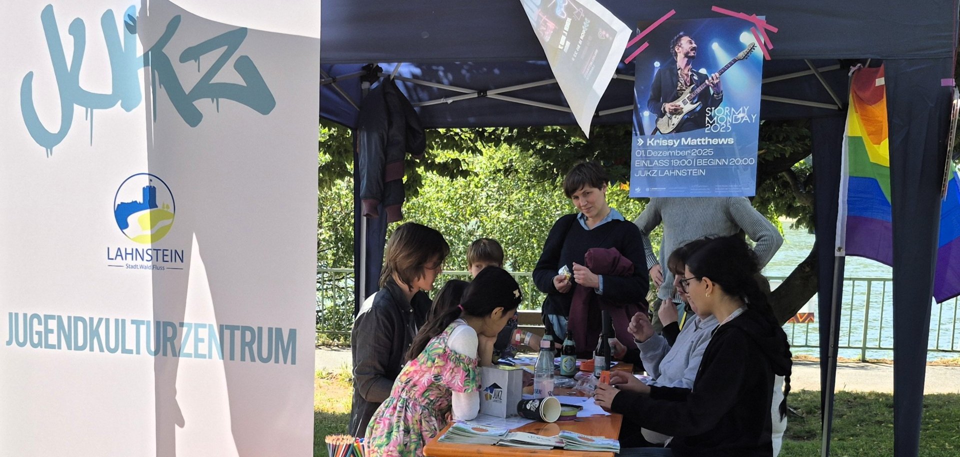 Unter einem Pavillon sitzen Kinder und Erwachsene an einem Tisch und basteln gemeinsam mit verschiedenen Materialien. Daneben steht ein Banner des Jugendkulturzentrums Lahnstein, im Hintergrund sind Bäume und der Rhein zu sehen.