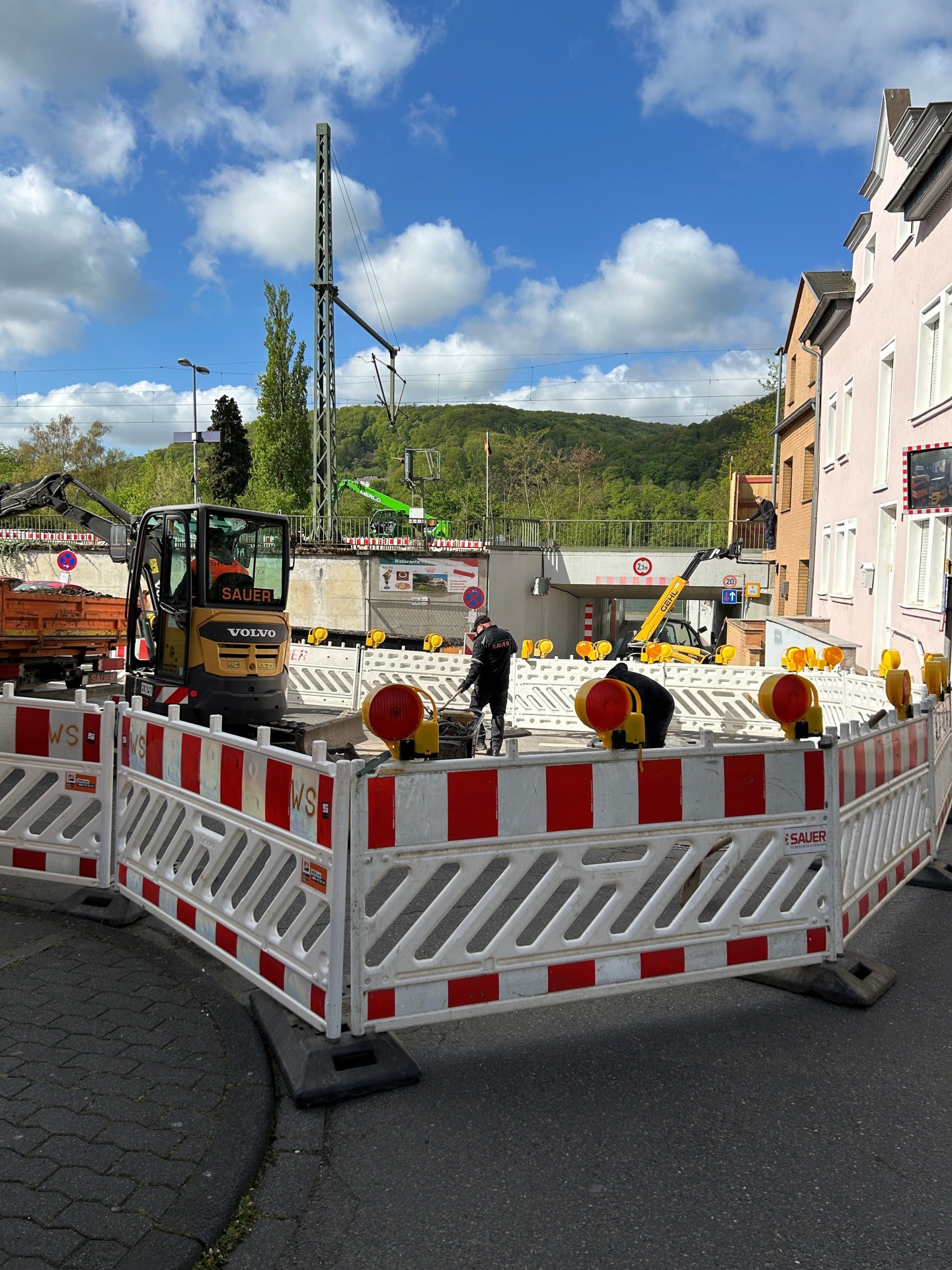 Baustelle an einer Straßenecke: Der Bereich ist mit rot-weißen Absperrgittern und Warnleuchten gesichert. Zwei Arbeiter sind innerhalb der Absperrung tätig, daneben steht ein kleiner Bagger. Im Hintergrund sind weitere Baugeräte, ein Bahnübergang bzw. eine Unterführung sowie Häuser und bewaldete Hügel unter blauem Himmel mit Wolken zu sehen.