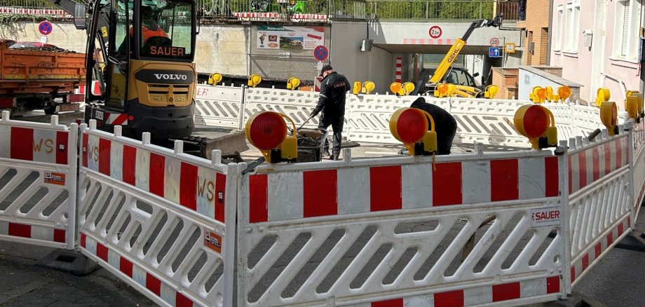 Baustelle an einer Straßenecke: Der Bereich ist mit rot-weißen Absperrgittern und Warnleuchten gesichert. Zwei Arbeiter sind innerhalb der Absperrung tätig, daneben steht ein kleiner Bagger. Im Hintergrund sind weitere Baugeräte, ein Bahnübergang bzw. eine Unterführung sowie Häuser und bewaldete Hügel unter blauem Himmel mit Wolken zu sehen.