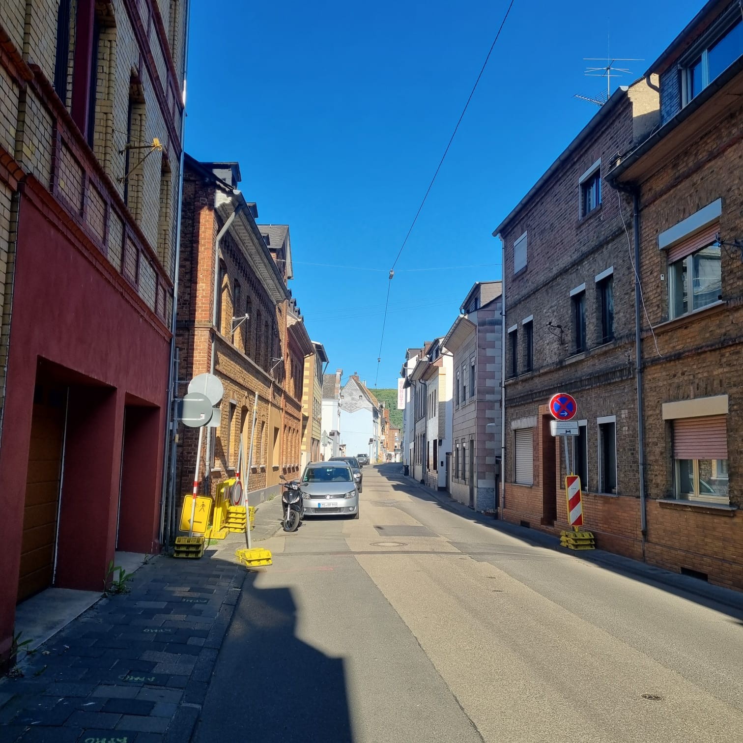 Schmale innerstädtische Straße mit beidseitiger Bebauung aus älteren Wohn- und Geschäftsgebäuden. Am Fahrbahnrand stehen ein geparktes Auto und ein Motorroller. Mehrere provisorische Verkehrsschilder und Absperrelemente deuten auf Bauarbeiten hin, darunter ein Halteverbotsschild und gelbe Warnbaken. Die Straße verläuft leicht ansteigend, im Hintergrund sind weitere Häuser und ein Hügel zu sehen, darüber ein klarer blauer Himmel.