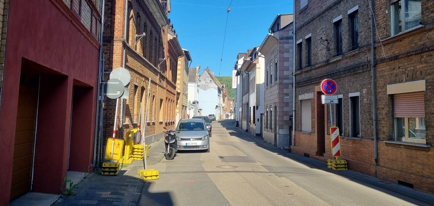 Schmale innerstädtische Straße mit beidseitiger Bebauung aus älteren Wohn- und Geschäftsgebäuden. Am Fahrbahnrand stehen ein geparktes Auto und ein Motorroller. Mehrere provisorische Verkehrsschilder und Absperrelemente deuten auf Bauarbeiten hin, darunter ein Halteverbotsschild und gelbe Warnbaken. Die Straße verläuft leicht ansteigend, im Hintergrund sind weitere Häuser und ein Hügel zu sehen, darüber ein klarer blauer Himmel.