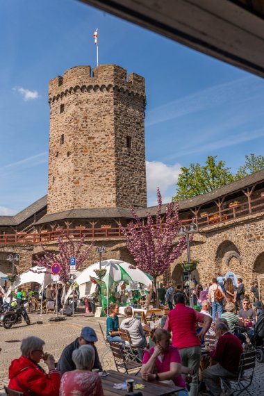 Eine weitläufige Außenaufnahme des belebten Salhofplatzes in Lahnstein bei strahlendem Sonnenschein. Im Hintergrund ragt der historische, steinerne Hexenturm mit Flagge empor, flankiert von einer mittelalterlichen Stadtmauer mit hölzernem Wehrgang. Auf dem Platz herrscht reges Treiben: Menschen sitzen an Holztischen, im Hintergrund sind weiße Eventzelte und blühende, rosafarbene Bäume zu sehen. Die Atmosphäre ist einladend und gemeinschaftlich.