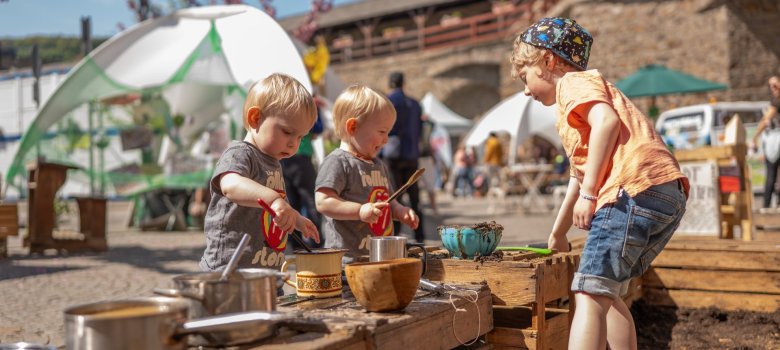 Drei kleine Kinder spielen konzentriert an einer provisorischen Außenküche aus rustikalen Holzkisten. Auf den Kisten stehen Töpfe, Tassen und Schüsseln, in denen mit Erde und Wasser „gematscht“ wird. Die Kinder tragen sommerliche Kleidung; im Hintergrund sind die hellen Zelte des Aktionstages und die historische Stadtmauer in Unschärfe zu sehen. Die Szene vermittelt spielerische Freude an der Natur.