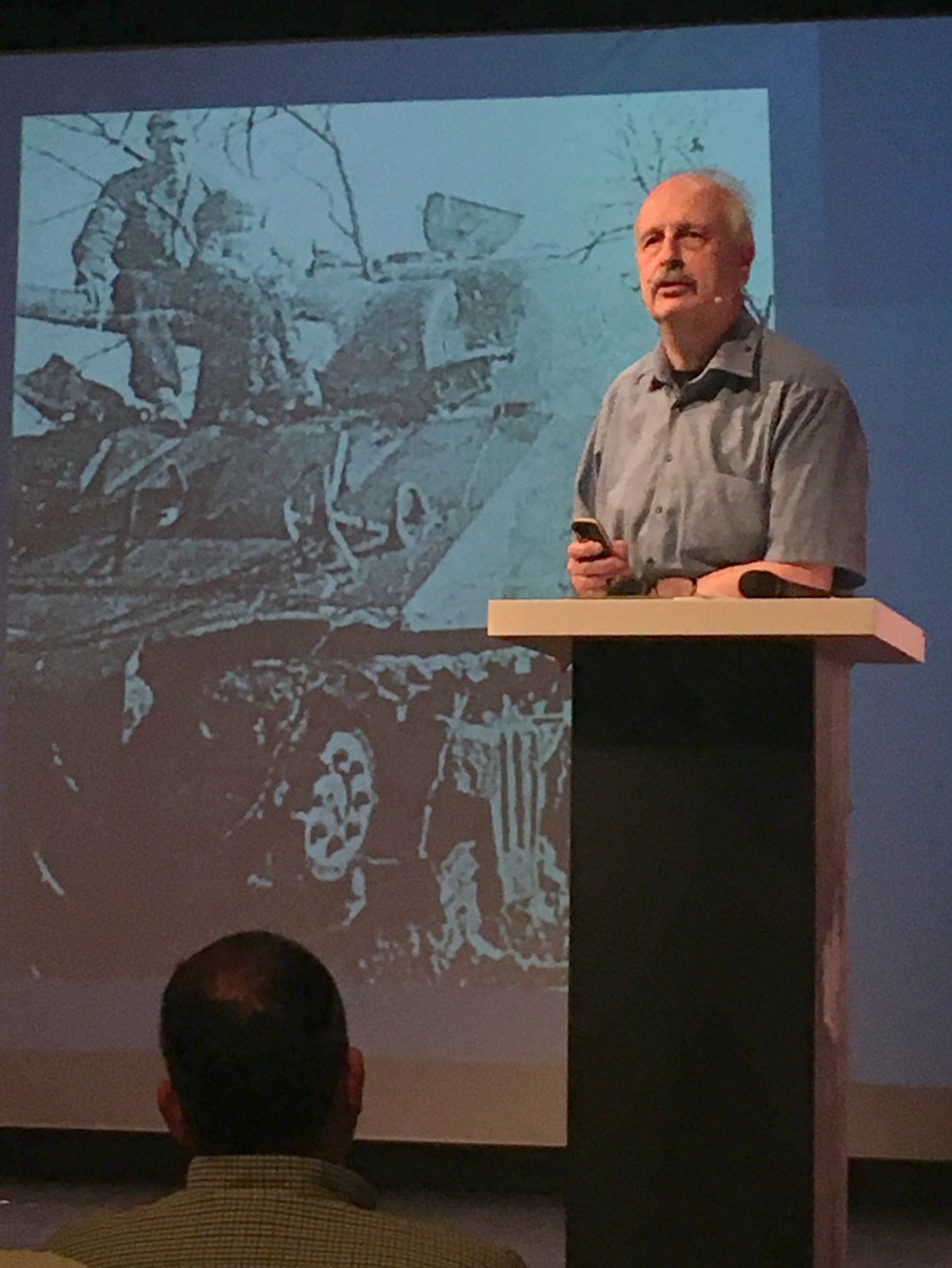 Ein Mann mit Schnurrbart steht an einem Rednerpult; im Hintergrund sieht man ein Schwarz-Weiß-Bild auf einer Leinwand