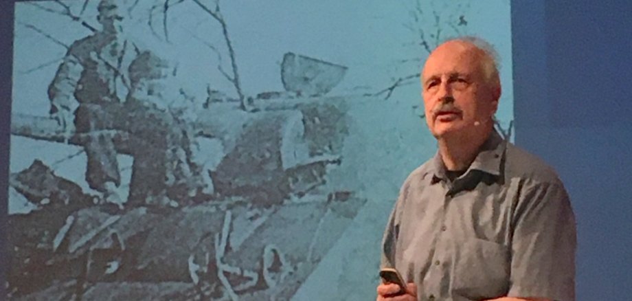 Ein Mann mit Schnurrbart steht an einem Rednerpult; im Hintergrund sieht man ein Schwarz-Weiß-Bild auf einer Leinwand