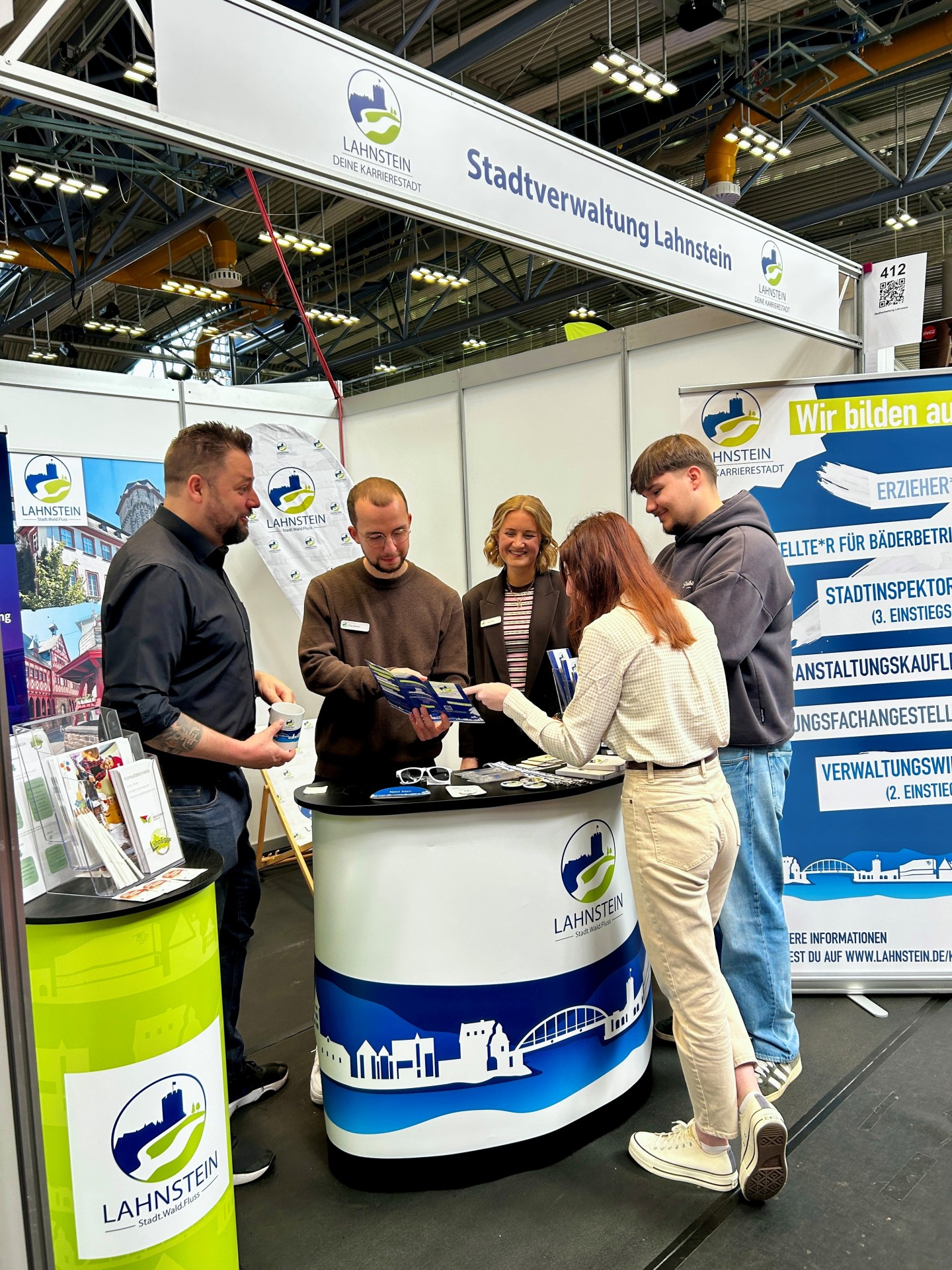 Fünf Personen stehen am Messestand der Stadtverwaltung Lahnstein in einer Ausstellungshalle und informieren sich gemeinsam über Ausbildungsangebote. Zwei Mitarbeiter erklären einer jungen Besucherin anhand von Flyern die Karrieremöglichkeiten, während weitere Beteiligte das Gespräch begleiten. Der Stand ist mit Lahnstein-Logos, Informationsmaterialien und einem großen Banner zu verschiedenen Ausbildungsberufen gestaltet.