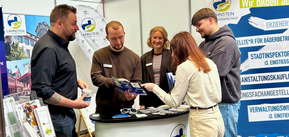 Fünf Personen stehen am Messestand der Stadtverwaltung Lahnstein in einer Ausstellungshalle und informieren sich gemeinsam über Ausbildungsangebote. Zwei Mitarbeiter erklären einer jungen Besucherin anhand von Flyern die Karrieremöglichkeiten, während weitere Beteiligte das Gespräch begleiten. Der Stand ist mit Lahnstein-Logos, Informationsmaterialien und einem großen Banner zu verschiedenen Ausbildungsberufen gestaltet.