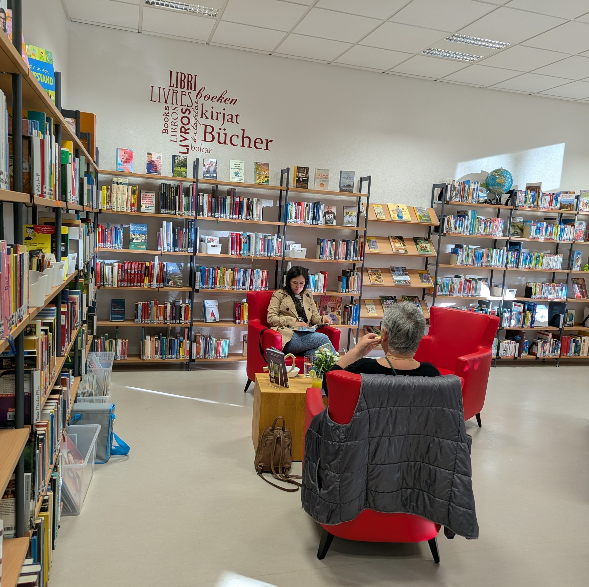 Innenansicht der Stadtbücherei Lahnstein während einer „Silent Reading“-Veranstaltung. Zwei Personen sitzen entspannt in leuchtend roten Sesseln zwischen gut gefüllten Bücherregalen und lesen. An der weißen Wand prangt eine dekorative Wortwolke, die das Wort „Bücher“ in verschiedenen Sprachen (u. a. Libri, Books, Livres) zeigt. Im Vordergrund steht ein kleiner Holztisch mit Getränken und Blumen; eine Tasche lehnt am Sessel. Die Atmosphäre wirkt hell, ruhig und einladend.