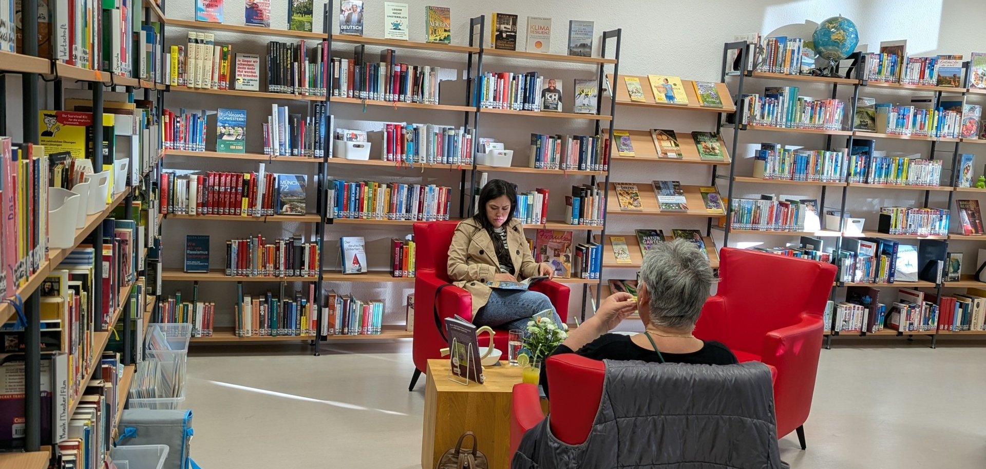 Innenansicht der Stadtbücherei Lahnstein während einer „Silent Reading“-Veranstaltung. Zwei Personen sitzen entspannt in leuchtend roten Sesseln zwischen gut gefüllten Bücherregalen und lesen. An der weißen Wand prangt eine dekorative Wortwolke, die das Wort „Bücher“ in verschiedenen Sprachen (u. a. Libri, Books, Livres) zeigt. Im Vordergrund steht ein kleiner Holztisch mit Getränken und Blumen; eine Tasche lehnt am Sessel. Die Atmosphäre wirkt hell, ruhig und einladend.