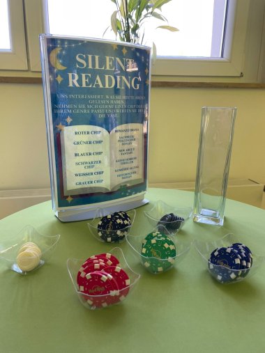 Nahaufnahme eines Tisches mit Informationen zum „Silent Reading“. Ein blaues Hinweisschild fordert die Gäste auf, einen farbigen Chip entsprechend ihrem gelesenen Genre in eine gläserne Vase zu werfen. Vor dem Schild stehen kleine Schalen mit farbigen Chips (weiß, rot, schwarz, grün, blau). Die Zuordnung lautet: Rot für Romanze, Grün für Sachbuch, Blau für Fantasy, Schwarz für Krimi, Weiß für Komödie und Grau für historische Romane. Im Hintergrund ist ein helles Fenster mit einer Pflanze zu sehen.