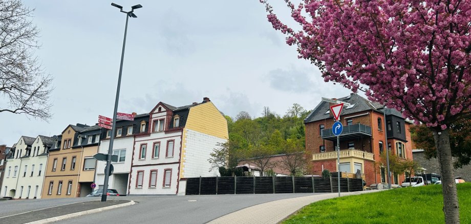 Blick bergauf in die Sebastianusstraße in Lahnstein an einem bewölkten Tag. Rechts im Vordergrund steht ein Baum mit üppigen rosa Blüten. An der ansteigenden Straße sind Autos vor einer Mischung aus modernen und historischen Stadthäusern. Ein Straßenschild weist den Weg nach rechts.