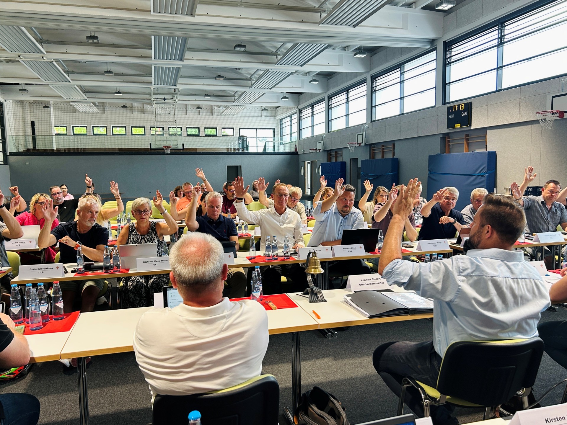 Mehrere Personen sitzen an langen Tischen in einer großen Sporthalle und heben die Hand für eine Abstimmung. Auf den Tischen stehen Wasserflaschen, Laptops, Unterlagen und Namensschilder. Im Vordergrund sitzen zwei Personen mit dem Rücken zur Kamera, eine davon hebt ebenfalls die Hand. Im Hintergrund sind hohe Fenster, Basketballkörbe und eine Anzeigetafel zu sehen.