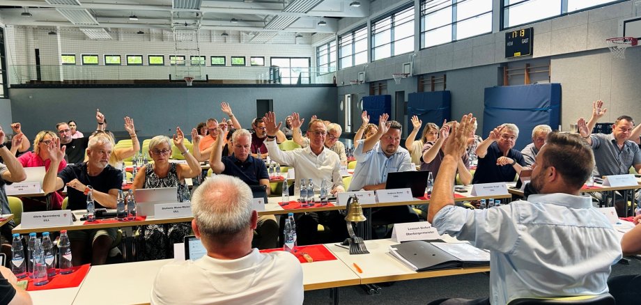 Stadtratssitzung Lahnstein Mehrere Personen sitzen an langen Tischen in einer großen Sporthalle und heben die Hand für eine Abstimmung. Auf den Tischen stehen Wasserflaschen, Laptops, Unterlagen und Namensschilder. Im Vordergrund sitzen zwei Personen mit dem Rücken zur Kamera, eine davon hebt ebenfalls die Hand. Im Hintergrund sind hohe Fenster, Basketballkörbe und eine Anzeigetafel zu sehen.