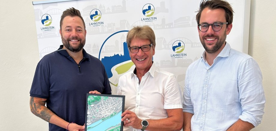 Drei Personen stehen vor einem Roll-up mit dem Logo der Stadt Lahnstein und lächeln in die Kamera. Die Person in der Mitte und die Person links halten gemeinsam ein gerahmtes Luftbild der Lahnsteiner Innenstadt mit dem Verlauf des Rheins in den Händen, das als Abschiedsgeschenk überreicht wird. Die Aufnahme entstand in einem hellen Büroraum und vermittelt den Eindruck einer feierlichen Verabschiedung.
