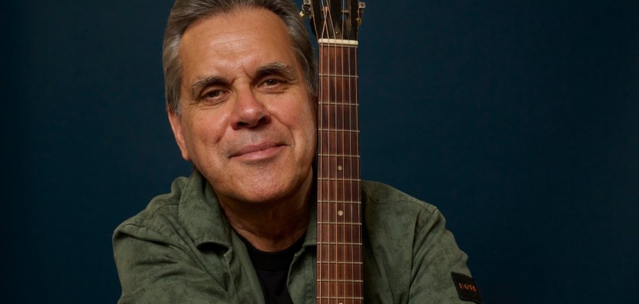 Biber Herrmann Porträt eines Mannes mit grauem Haar vor dunkelblauem Hintergrund. Er sitzt mit verschränkten Armen und lehnt sich an eine Gitarre mit metallischer Decke. Der Musiker trägt ein olivgrünes Hemd über einem dunklen Shirt und blickt leicht lächelnd direkt in die Kamera. Die Aufnahme wirkt ruhig, konzentriert und professionell inszeniert.