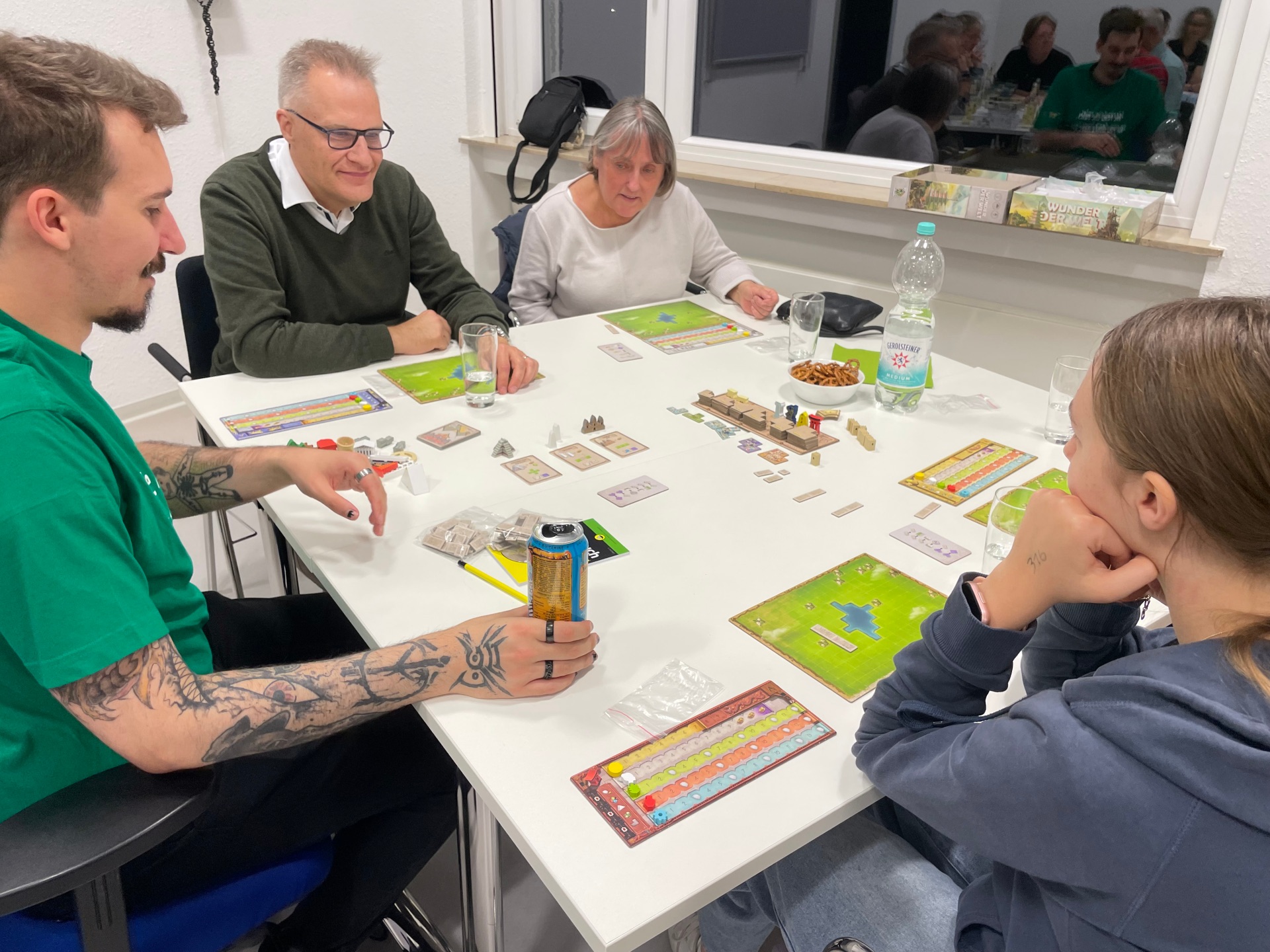 Fünf Personen sitzen an einem Tisch und spielen gemeinsam ein Brettspiel. Auf dem Tisch liegen Spielpläne, Karten, Spielfiguren und weiteres Spielmaterial sowie Getränke und eine Schale mit Salzgebäck. Die Teilnehmenden konzentrieren sich auf das Spielgeschehen und unterhalten sich. Im Hintergrund spiegelt sich eine weitere Spielgruppe in der Fensterscheibe.