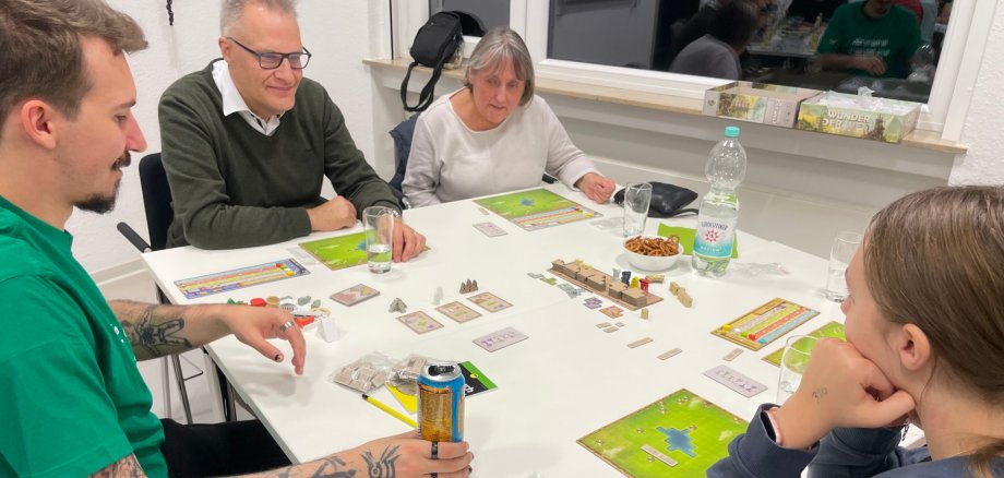 Spieleabend Stadtbücherei Lahnstein Fünf Personen sitzen an einem Tisch und spielen gemeinsam ein Brettspiel. Auf dem Tisch liegen Spielpläne, Karten, Spielfiguren und weiteres Spielmaterial sowie Getränke und eine Schale mit Salzgebäck. Die Teilnehmenden konzentrieren sich auf das Spielgeschehen und unterhalten sich. Im Hintergrund spiegelt sich eine weitere Spielgruppe in der Fensterscheibe.