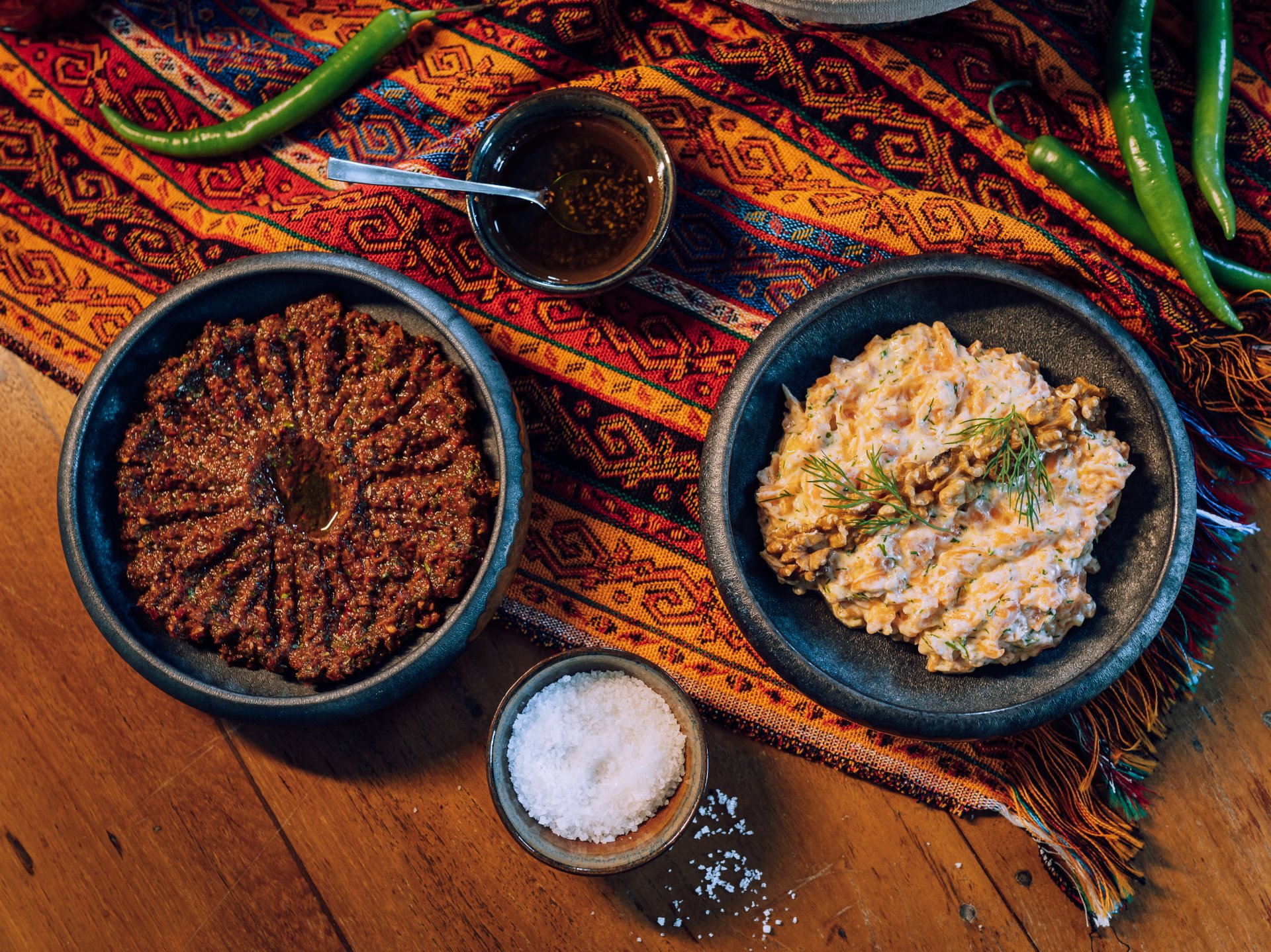 Zwei Schalen mit orientalischen Speisen stehen auf einem bunt gemusterten Tuch auf einem Holztisch. Die linke Schale enthält einen dunkelroten Dip mit Öl in der Mitte, die rechte einen cremigen Salat, garniert mit Walnüssen und frischem Dill. Daneben liegen grüne Chilischoten sowie kleine Schälchen mit Gewürzen und grobem Salz.