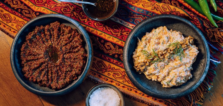 Zwei Schalen mit orientalischen Speisen stehen auf einem bunt gemusterten Tuch auf einem Holztisch. Die linke Schale enthält einen dunkelroten Dip mit Öl in der Mitte, die rechte einen cremigen Salat, garniert mit Walnüssen und frischem Dill. Daneben liegen grüne Chilischoten sowie kleine Schälchen mit Gewürzen und grobem Salz.