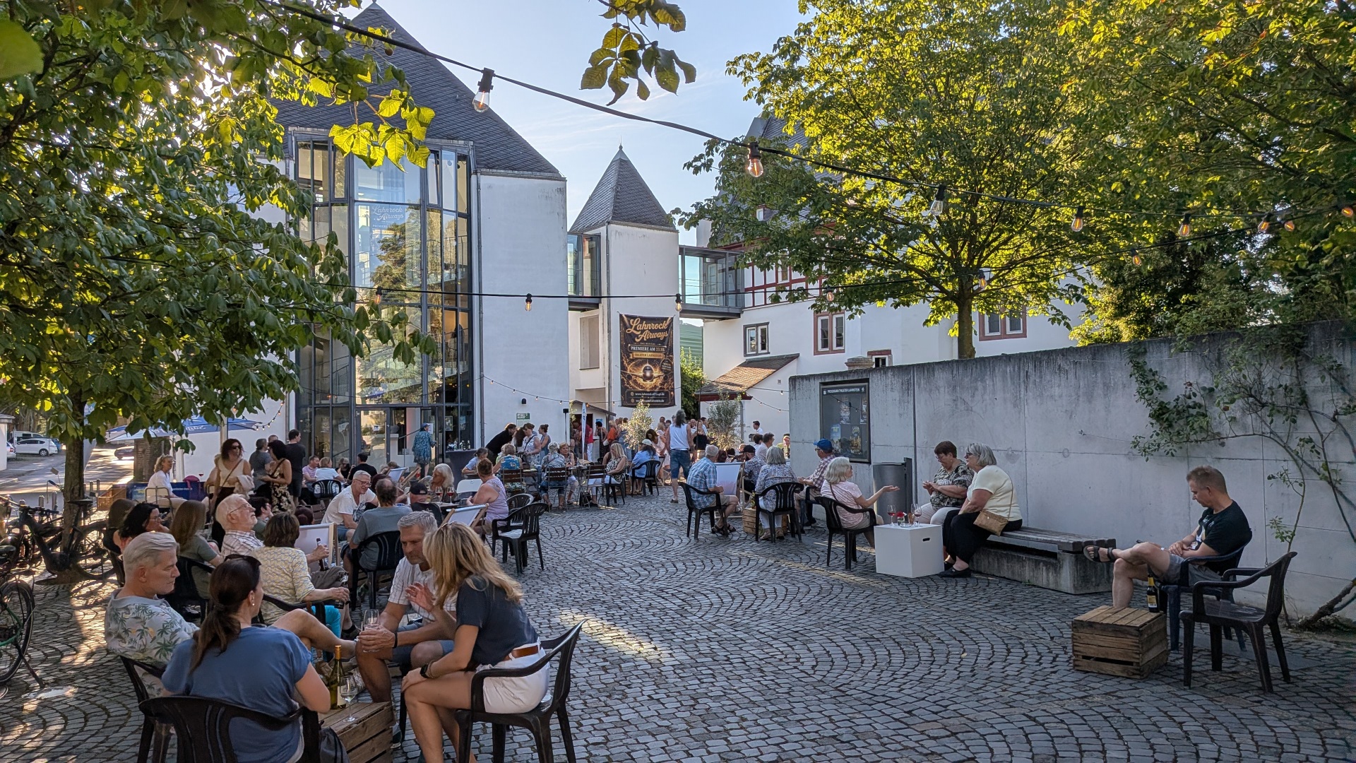 Auf einem gepflasterten Innenhof sitzen zahlreiche Menschen an Tischen und genießen einen sommerlichen Abend. Zwischen den Bäumen sind Lichterketten gespannt und schaffen eine stimmungsvolle Atmosphäre. Im Hintergrund verbindet ein moderner Glasbau historische Gebäude. Ein großes Banner kündigt den „Lahnrock Always“-Abend an. Die Gäste unterhalten sich, trinken und verbringen gemeinsam Zeit im Freien.