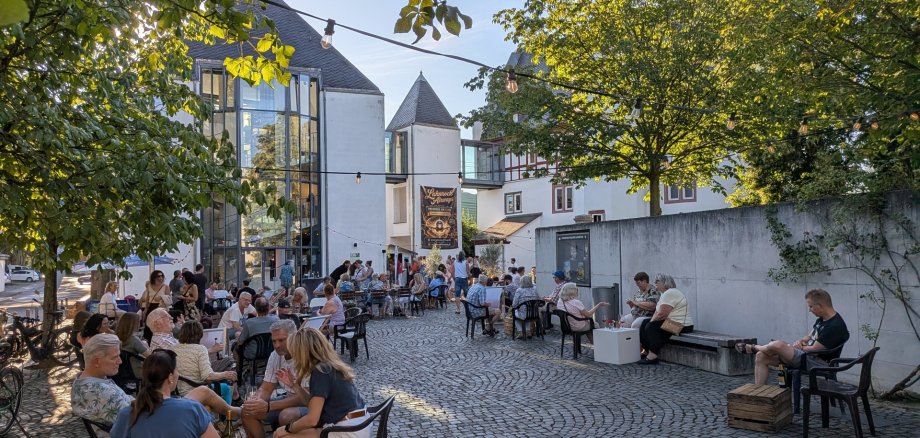 Lahnsteiner Treff Auf einem gepflasterten Innenhof sitzen zahlreiche Menschen an Tischen und genießen einen sommerlichen Abend. Zwischen den Bäumen sind Lichterketten gespannt und schaffen eine stimmungsvolle Atmosphäre. Im Hintergrund verbindet ein moderner Glasbau historische Gebäude. Ein großes Banner kündigt den „Lahnrock Always“-Abend an. Die Gäste unterhalten sich, trinken und verbringen gemeinsam Zeit im Freien.