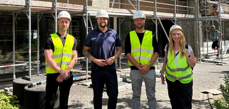 Vier Personen tragen Schutzhelme und Warnwesten und stehen vor einem mehrgeschossigen Gebäude, das vollständig von einem Baugerüst umgeben ist. Hinter ihnen arbeiten Handwerker an der Fassade. Das Gebäude befindet sich in einer fortgeschrittenen Sanierungs- oder Bauphase, Fenster sind bereits eingebaut. Die Gruppe blickt in die Kamera und dokumentiert den Baufortschritt.