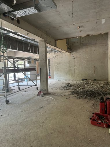 Innenansicht einer Baustelle mit freigelegten Betonwänden und -decken. Ein Baugerüst steht im linken Bereich, von der Decke hängen Kabel und Installationsleitungen. Rechts liegen Bauschutt und Metallreste auf dem Boden, daneben Werkzeugkoffer. Der Raum befindet sich im Rohbau und wird für den weiteren Innenausbau vorbereitet.