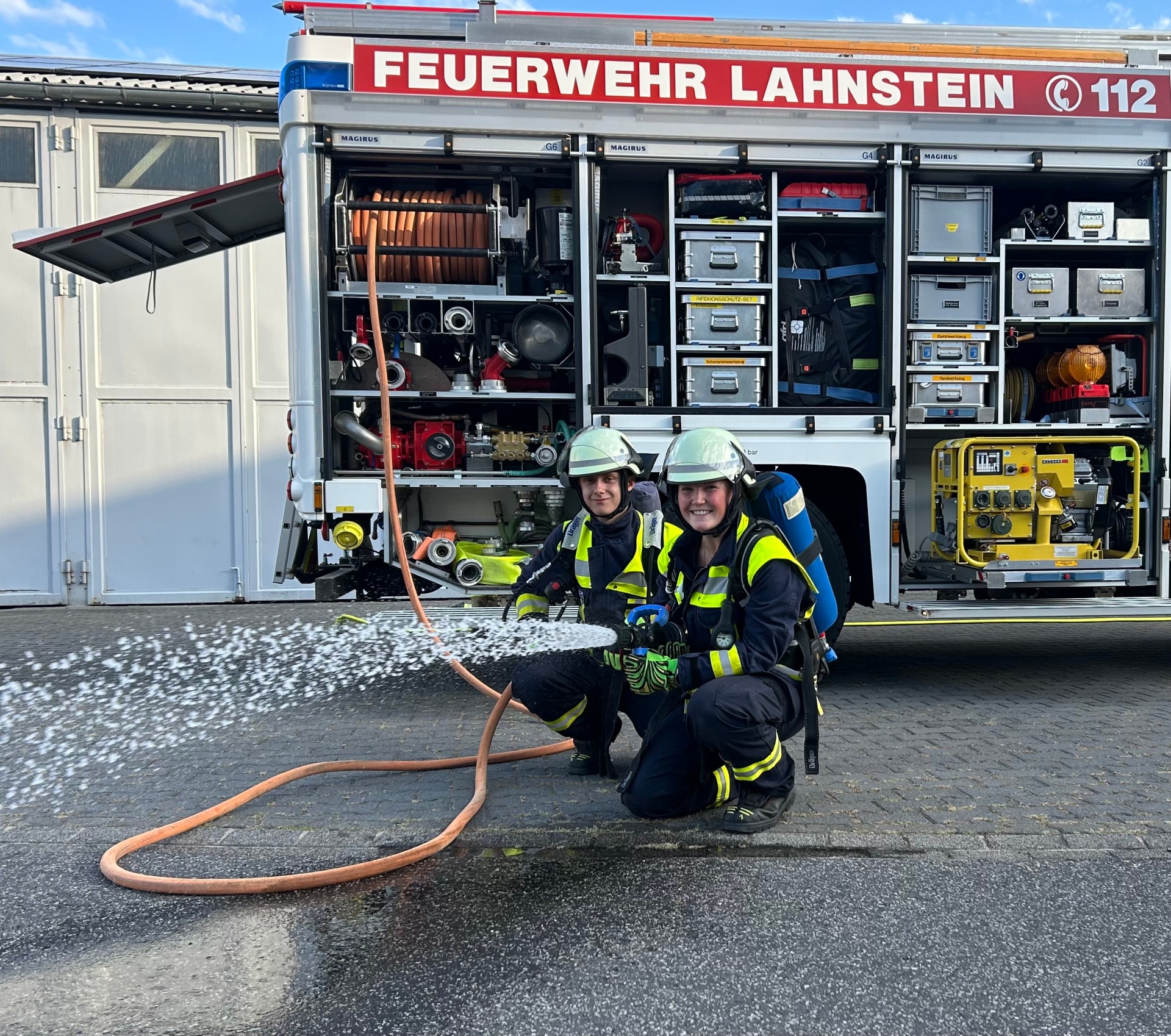 Zwei Einsatzkräfte der Feuerwehr Lahnstein knien in vollständiger Schutzkleidung mit Helmen und Atemschutzgeräten vor einem geöffneten Feuerwehrfahrzeug. Eine der Personen hält ein Strahlrohr, aus dem Wasser spritzt. In den geöffneten Gerätefächern des Fahrzeugs sind Schläuche, Pumpen und weitere Feuerwehrausrüstung zu sehen.