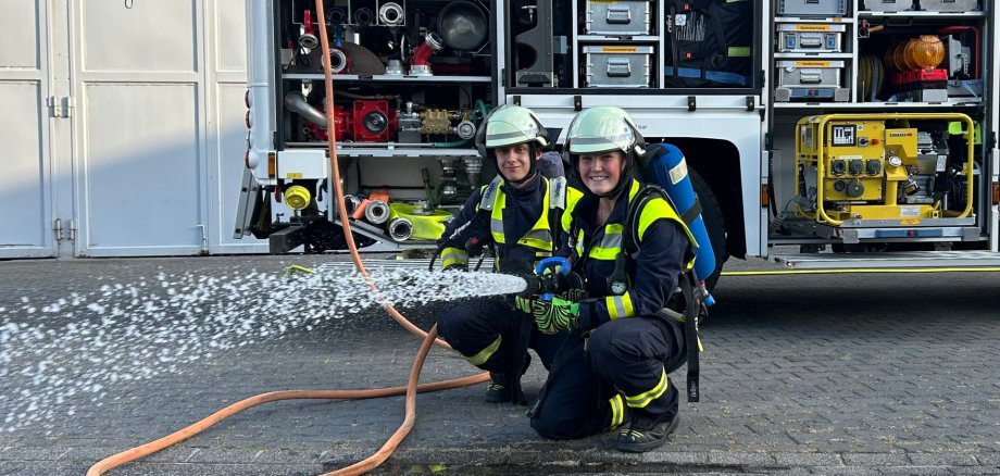 Zwei Einsatzkräfte der Feuerwehr Lahnstein knien in vollständiger Schutzkleidung mit Helmen und Atemschutzgeräten vor einem geöffneten Feuerwehrfahrzeug. Eine der Personen hält ein Strahlrohr, aus dem Wasser spritzt. In den geöffneten Gerätefächern des Fahrzeugs sind Schläuche, Pumpen und weitere Feuerwehrausrüstung zu sehen.