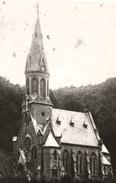Simultankirche Friedrichssegten, 1910 Historische Schwarz-Weiß-Aufnahme der ehemaligen Simultankirche in Friedrichssegen vor ihrem Abriss. Zu sehen ist das neugotische Kirchengebäude mit hohem, spitzem Kirchturm, Rundfenster und hohen Kirchenfenstern vor einem bewaldeten Hang.