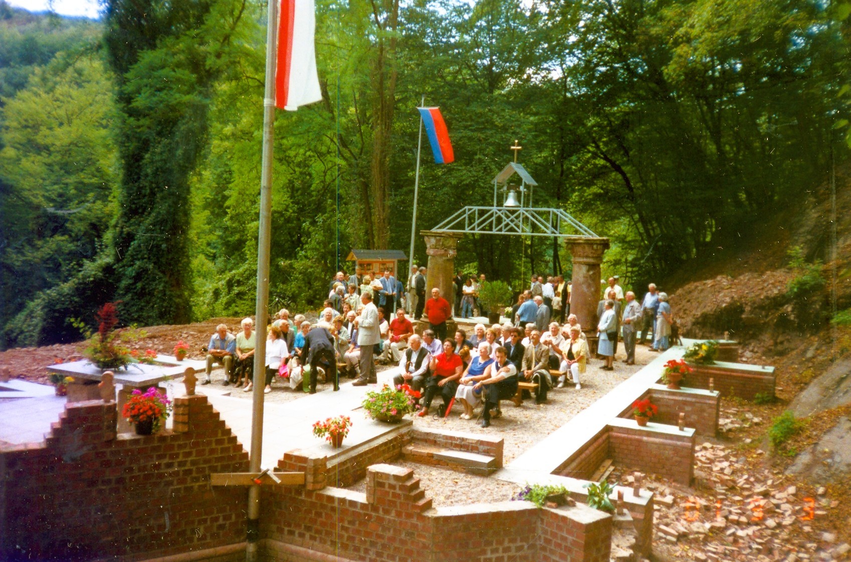 Historische Farbaufnahme der freigelegten Überreste der ehemaligen Simultankirche in Friedrichssegen. Zahlreiche Menschen sitzen und stehen auf dem von Wald umgebenen Gelände zwischen erhaltenen Mauerteilen und Säulen. Im Hintergrund befindet sich eine Konstruktion mit einer Glocke und einem Kreuz; das Gelände ist mit Blumen geschmückt.