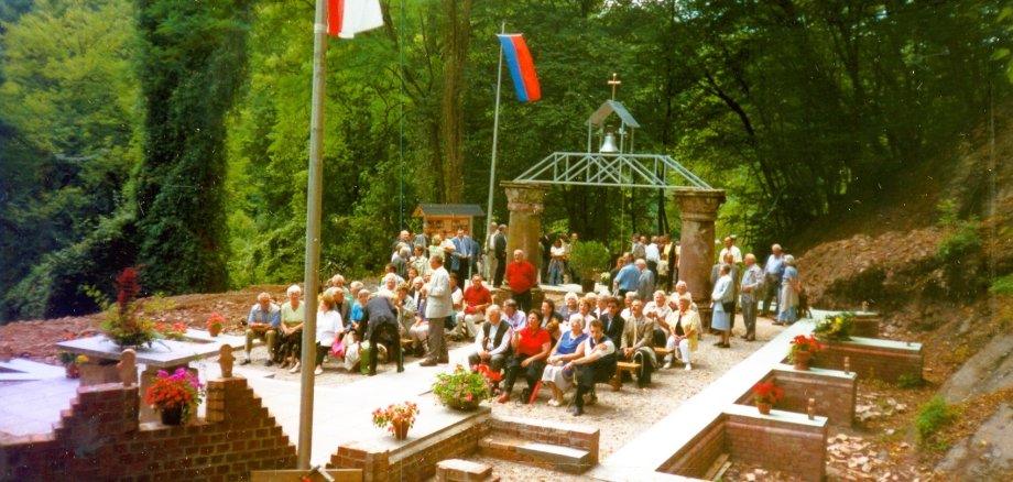 Simultankirche in Friedrichssegen Historische Farbaufnahme der freigelegten Überreste der ehemaligen Simultankirche in Friedrichssegen. Zahlreiche Menschen sitzen und stehen auf dem von Wald umgebenen Gelände zwischen erhaltenen Mauerteilen und Säulen. Im Hintergrund befindet sich eine Konstruktion mit einer Glocke und einem Kreuz; das Gelände ist mit Blumen geschmückt.