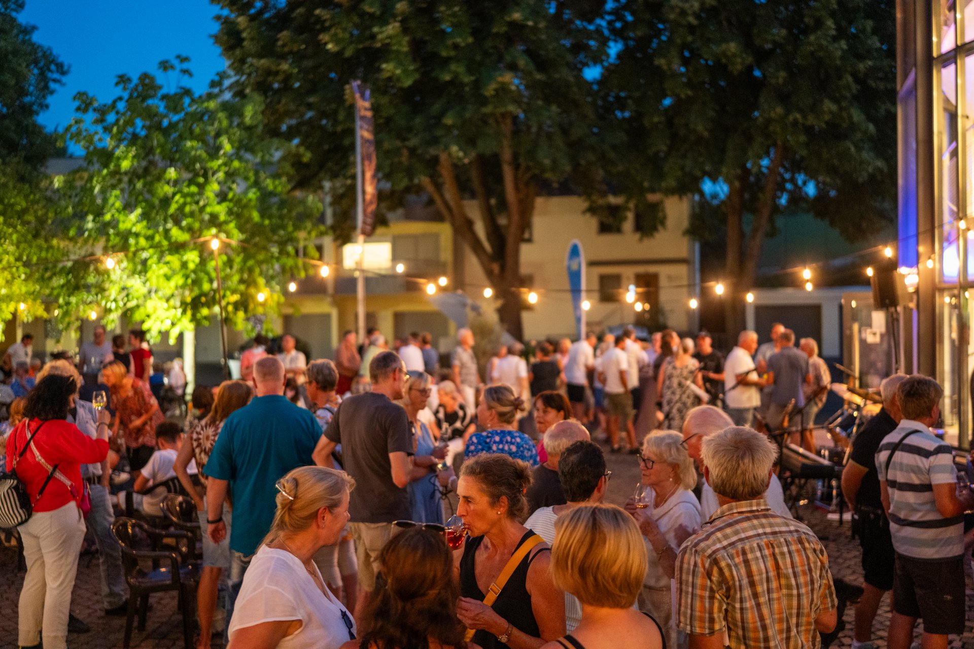 Zahlreiche Menschen feiern am Abend bei einem Straßenfest unter Lichterketten. Die Gäste stehen in Gruppen zusammen oder sitzen an Tischen; Bäume und warmes Licht sorgen für eine sommerliche Atmosphäre.