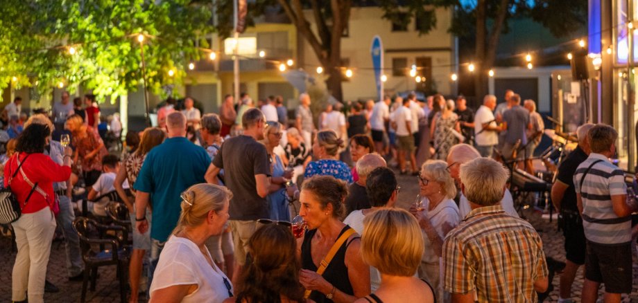 Zahlreiche Menschen feiern am Abend bei einem Straßenfest unter Lichterketten. Die Gäste stehen in Gruppen zusammen oder sitzen an Tischen; Bäume und warmes Licht sorgen für eine sommerliche Atmosphäre.