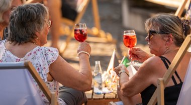 Zwei Frauen sitzen bei Sonnenschein in Liegestühlen und stoßen mit Gläsern eines roten Getränks an. Zwischen ihnen stehen Getränke und kleine italienische Fähnchen auf einem Tisch.