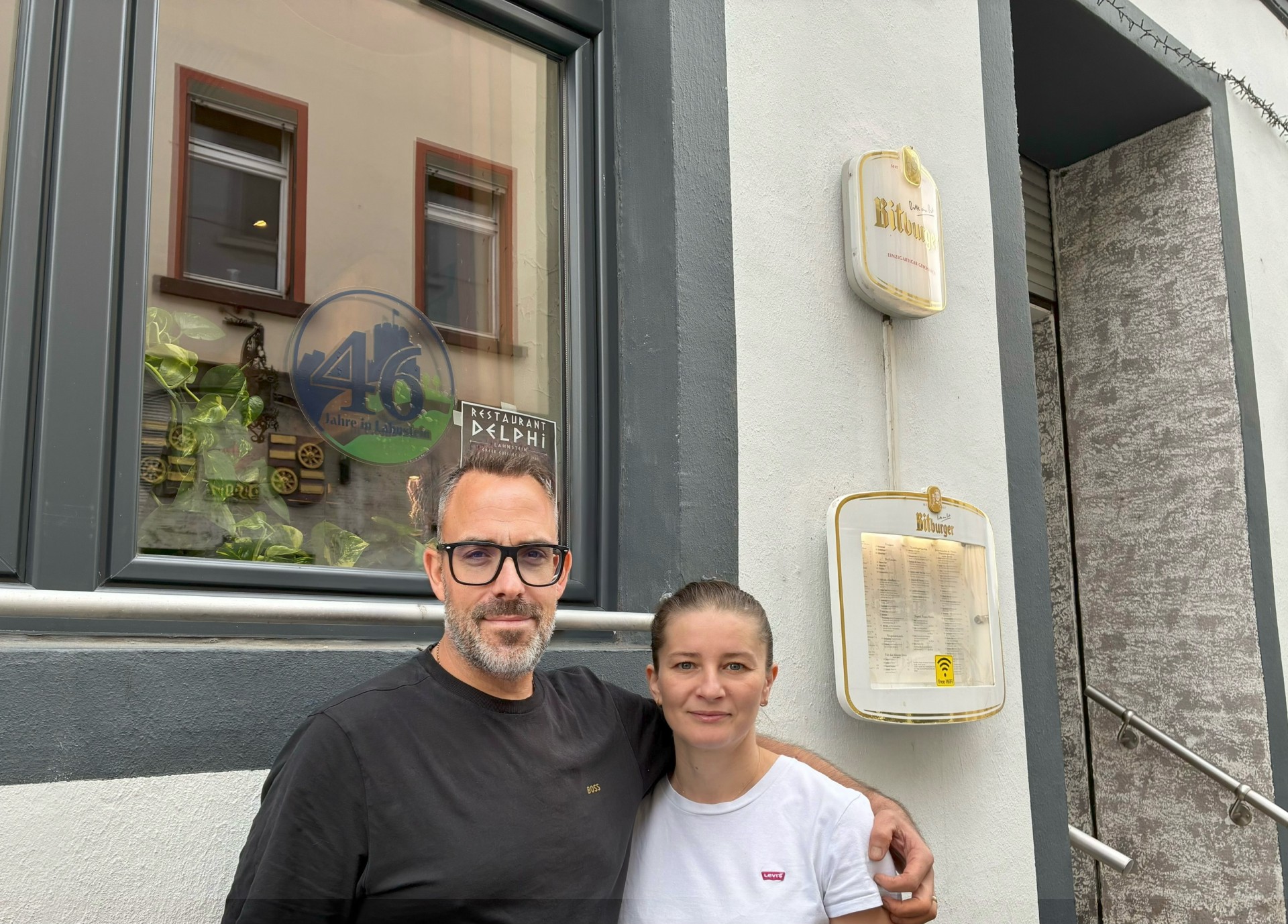 Ein Mann und eine Frau stehen nebeneinander vor der Fassade eines Restaurants und blicken in die Kamera. Der Mann trägt eine schwarze Brille und hat einen Arm um die Frau gelegt. Hinter ihnen ist ein Fenster zu sehen, in dem unter anderem ein Aufkleber mit der Aufschrift „Restaurant Delphi“ angebracht ist. Rechts neben den beiden hängen ein Bitburger-Schild und ein Schaukasten mit der Speisekarte an der Hauswand.